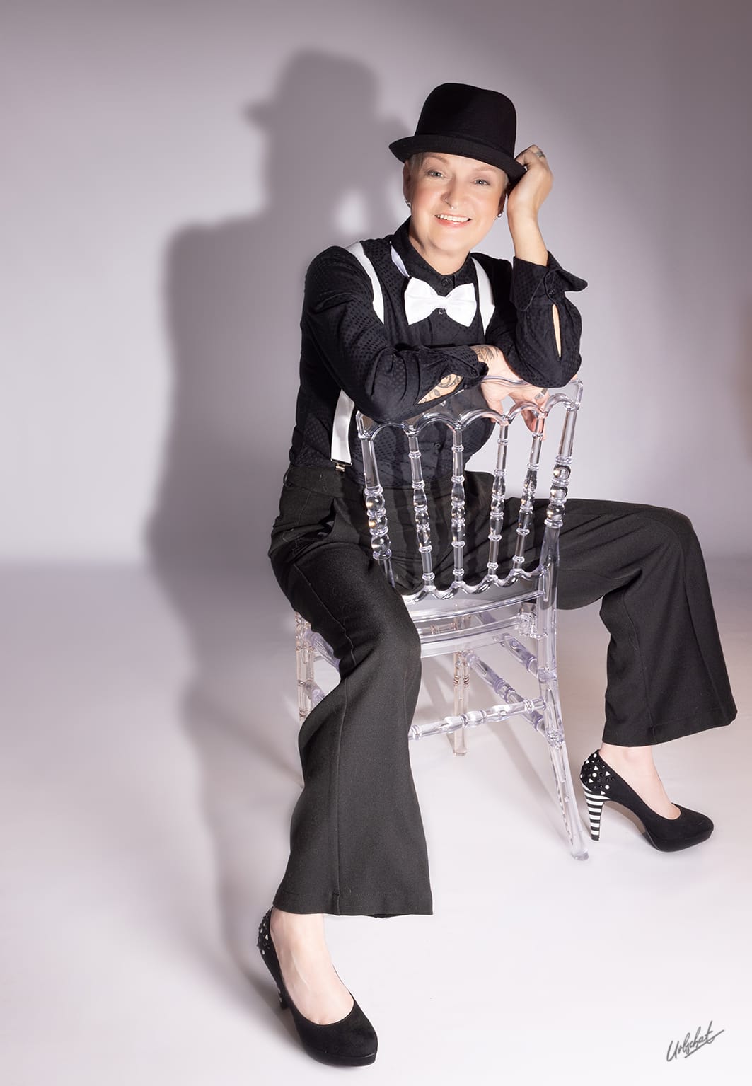 Katlui — Character look: cabaret tuxedo with black fedora, white suspenders and bow tie, seated on transparent acrylic chair